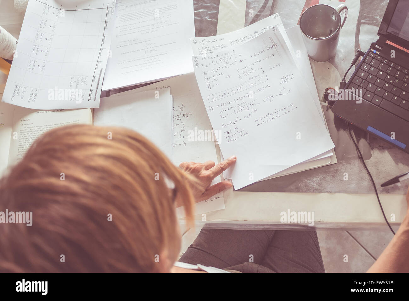Femme assise au bureau en face de l'ordinateur portable, lecture mathématiques formule sur papier blanc. Mug vide et de nombreux documents épars. Banque D'Images