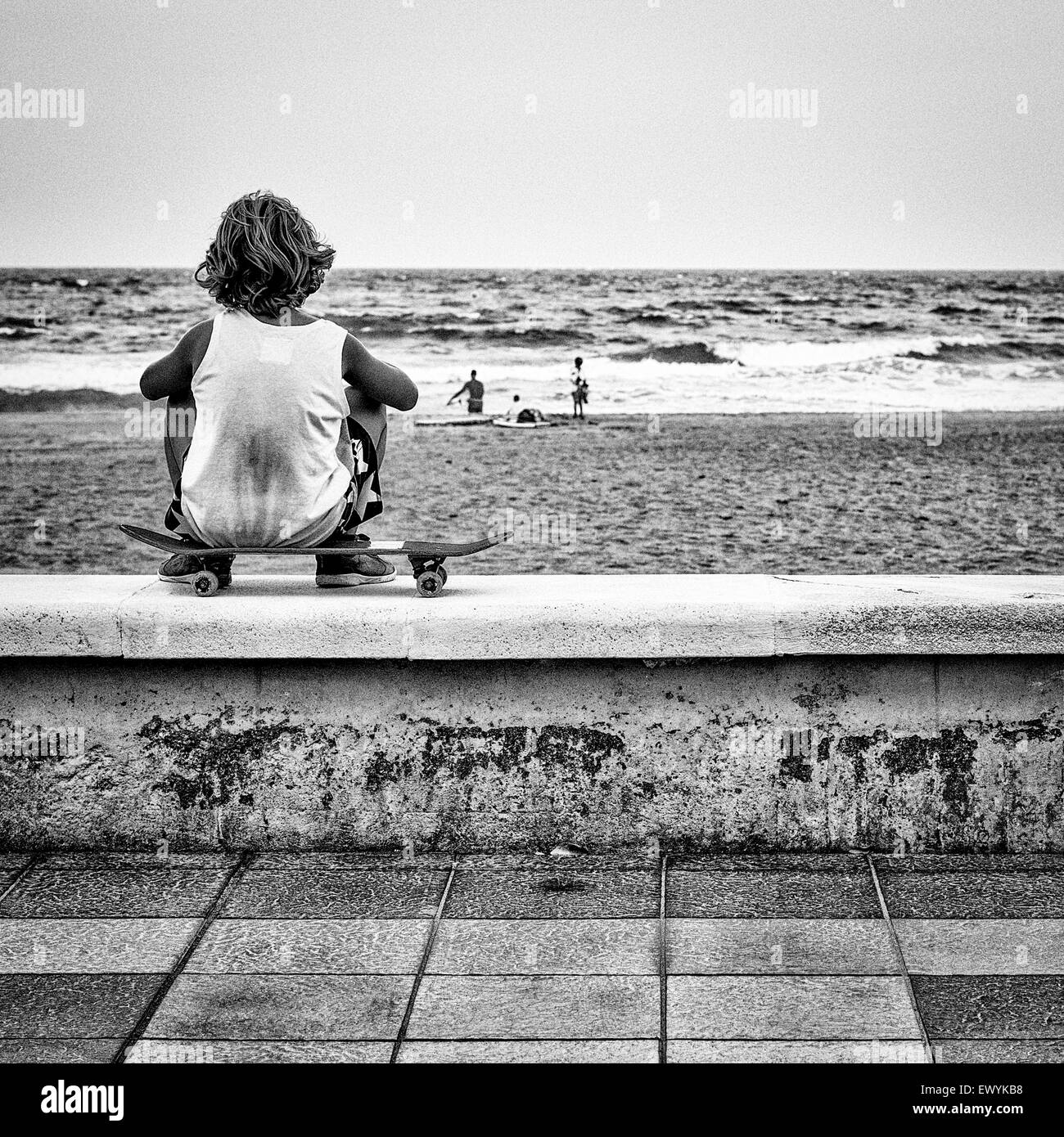 Un garçon assis sur sa planche à roulettes de la mer est à regarder les surfeurs se préparer est représentée dans cette photo en noir et blanc Banque D'Images