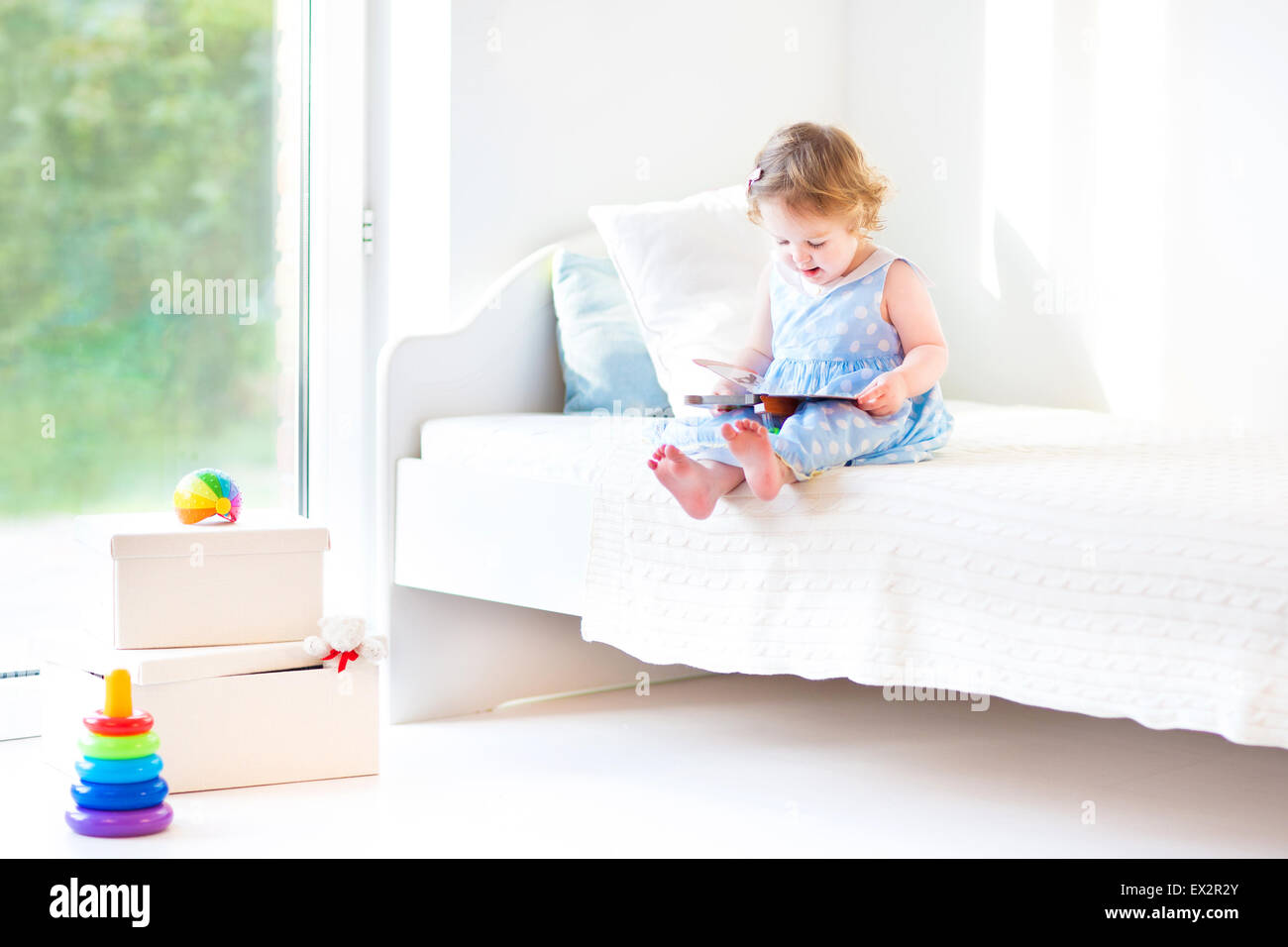 Adorable bébé fille lire un livre assis sur un lit blanc dans une grande fenêtre avec vue sur le jardin Banque D'Images