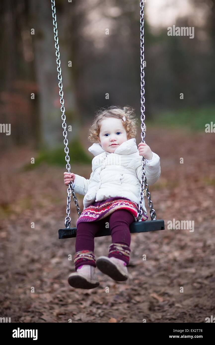 Adorable bébé fille avec des cheveux bouclés portant une veste chaude et blanche robe violette s'amusant sur une balançoire dans un parc automne sombre Banque D'Images