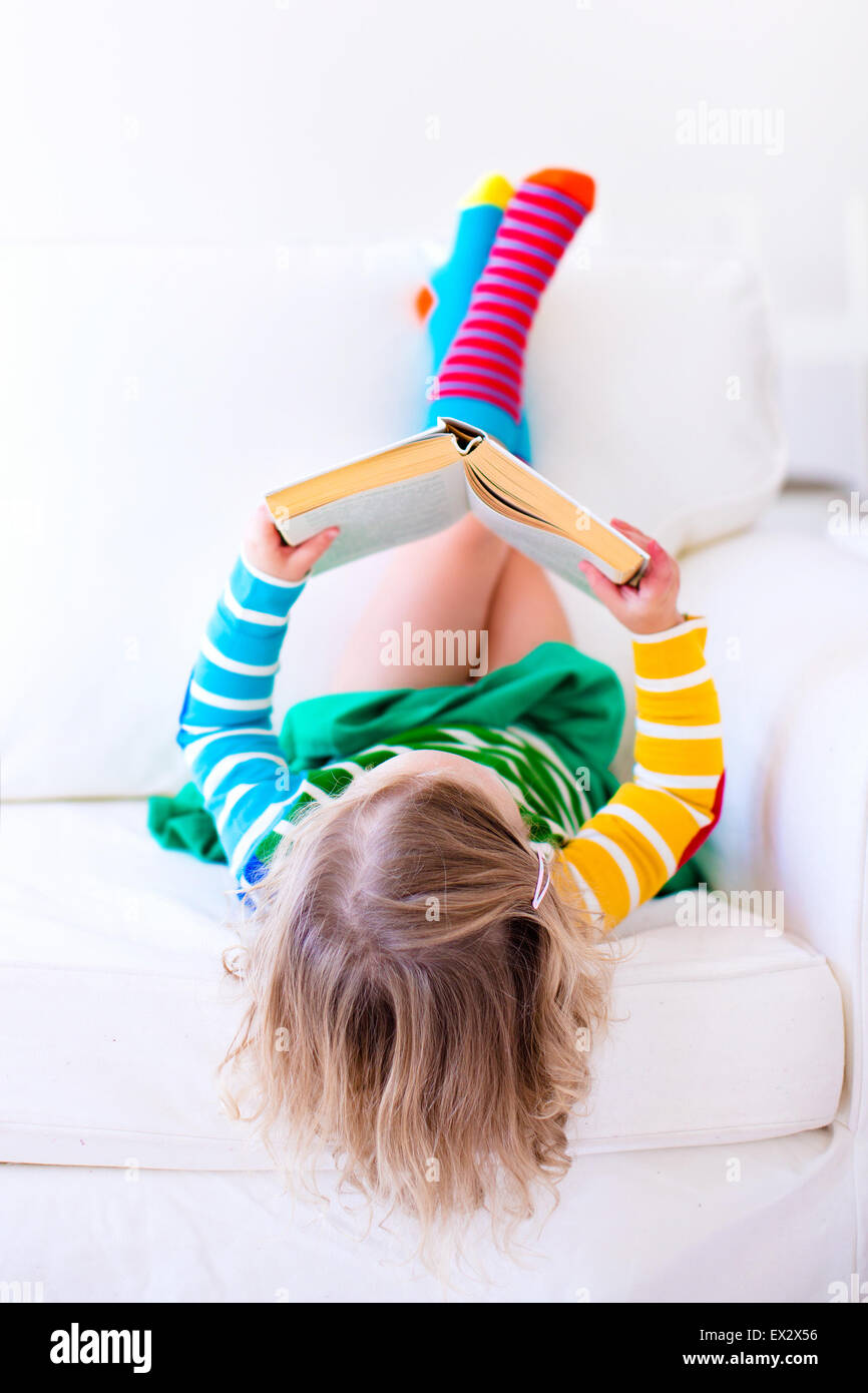 Petite fille lire un livre de vous détendre sur un canapé blanc. Lire des livres pour enfants à la maison ou à la garderie. L'apprentissage et les enfants à faire leurs devoirs Banque D'Images