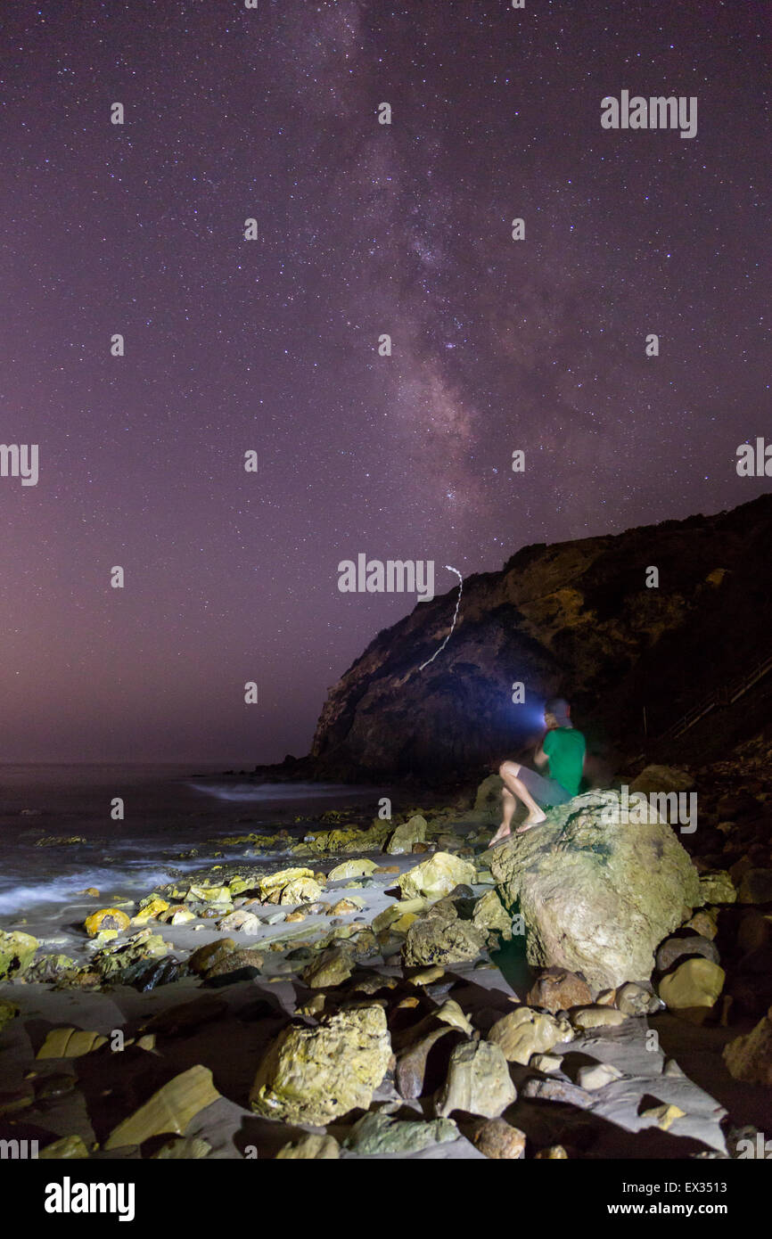 Un homme est assis sur un rocher à Malibu pointant sa lampe de projecteur pour les étoiles et Milky Way. Banque D'Images