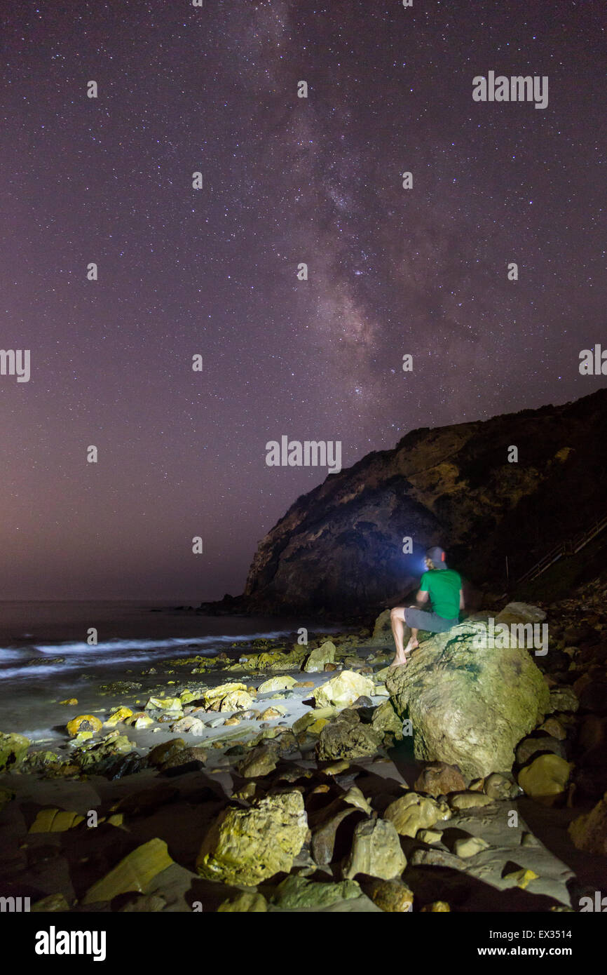 Un homme est assis sur un rocher à Malibu pointant sa lampe de projecteur pour les étoiles et Milky Way. Banque D'Images