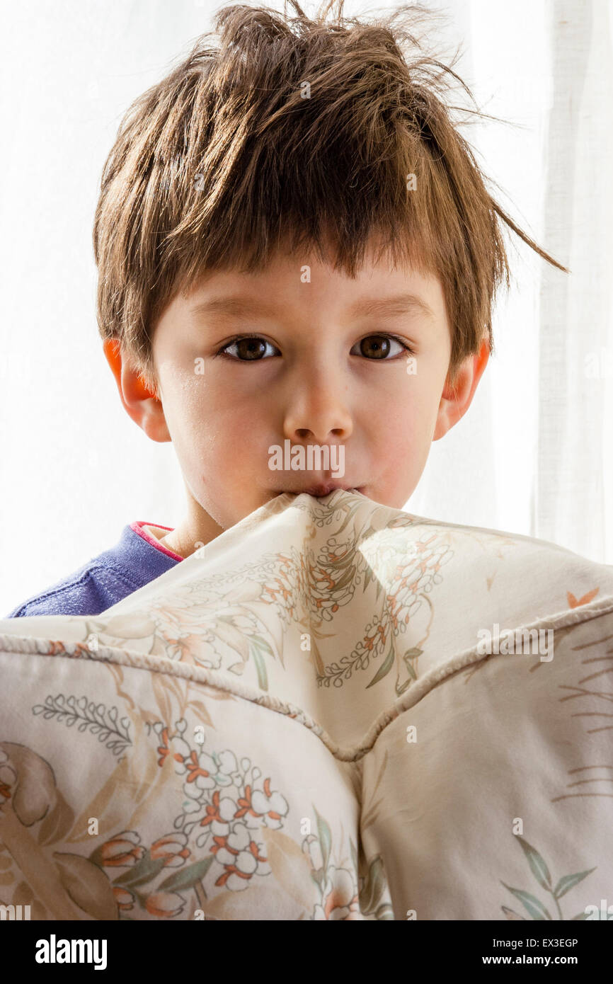 Enfant, 6-7 ans, face à la tenue par ses dents. Eye-contact, Caucasian boy. Tête et épaules. À l'intérieur avec un rideau lumineux arrière-plan. Banque D'Images