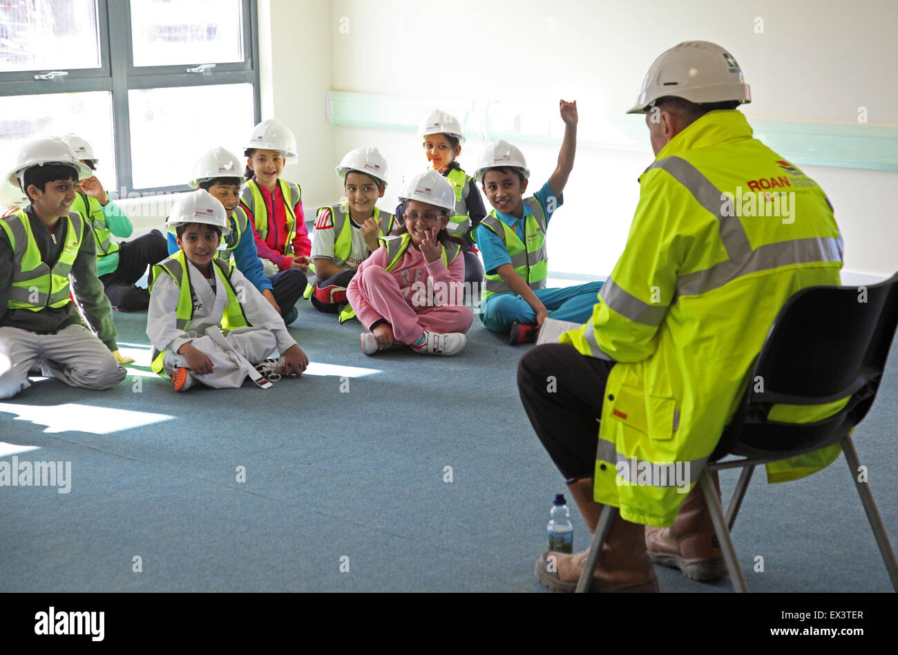 Les jeunes enfants de l'école des casques de sécurité et des vestes haute vis sur un site visite à une nouvelle extension de leur école. L'élève lève la main pour répondre à une question Banque D'Images