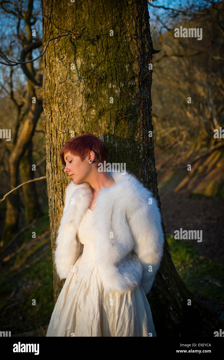 Contes modernes : une jeune femme androgyne caucasienne girl alone par  elle-même dans une forêt bois appuyé contre un arbre portant une robe de  mariée blanche et veste en fourrure Photo Stock -