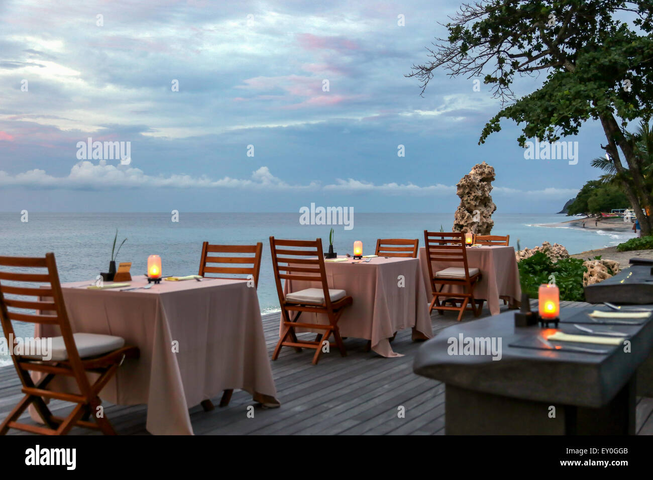 Quatre tables de restaurant sur le plancher en bois à côté de la plage pendant le coucher du soleil. Chaque table dispose de deux chaises et d'une lampe sur la mer. Banque D'Images