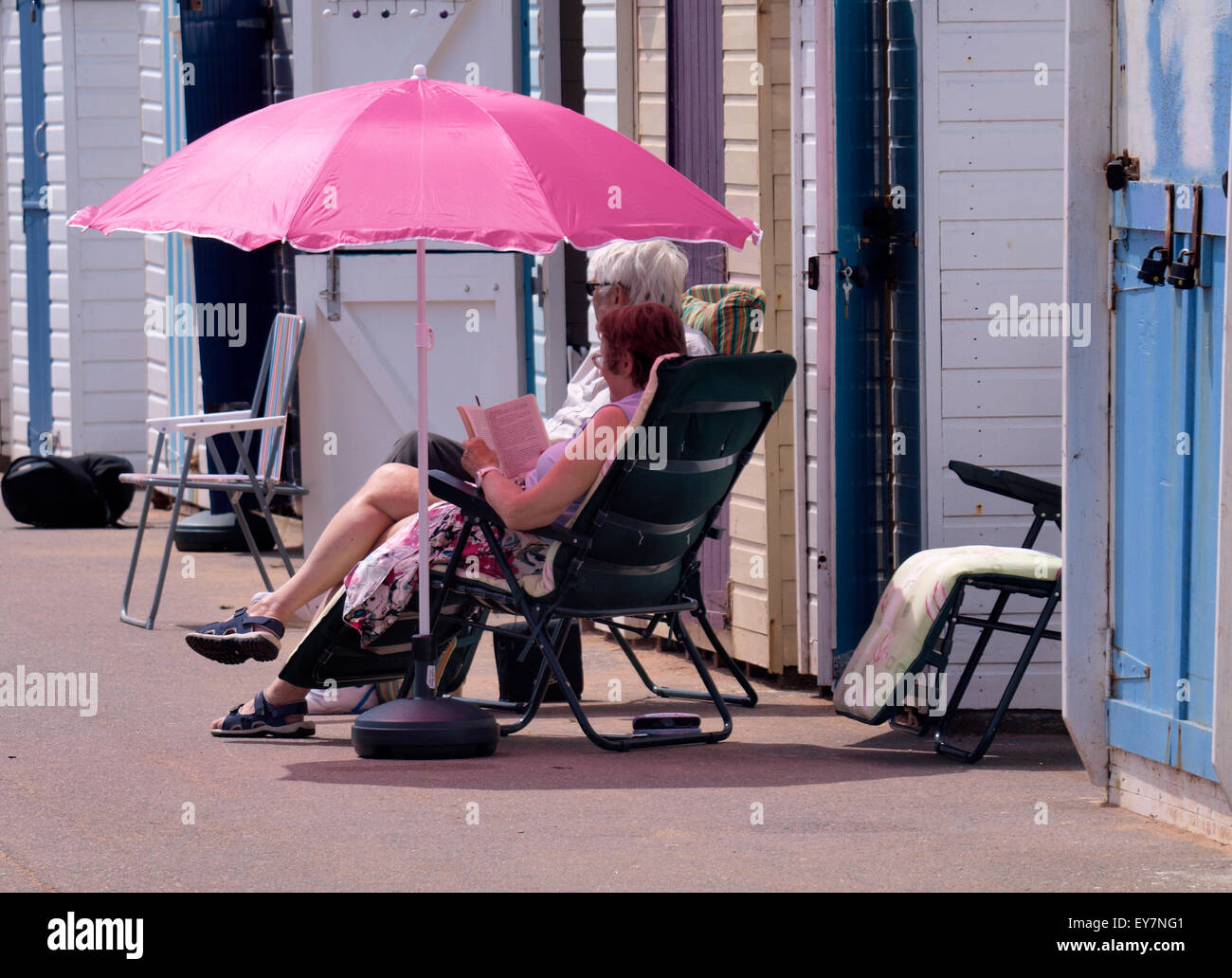 Couple assis sous un parasol rose en face de cabanes de plage de Goodrington Sands Paignton, Devon, UK Banque D'Images