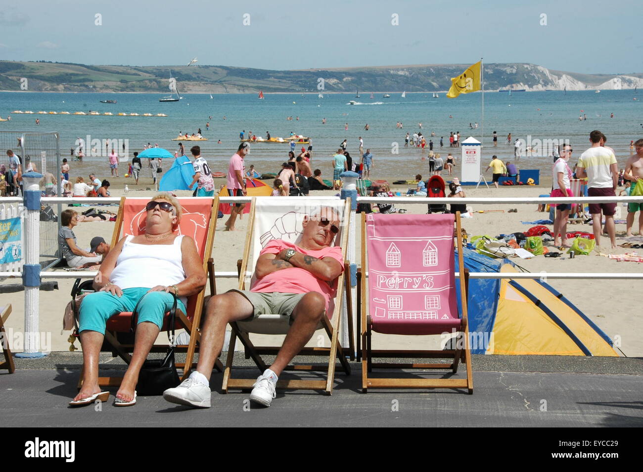 Par un beau jour d'été sur l'Esplanade de Weymouth, deux personnes répéter sur des chaises longues avec un fond d'animation de l'activité balnéaire. Banque D'Images