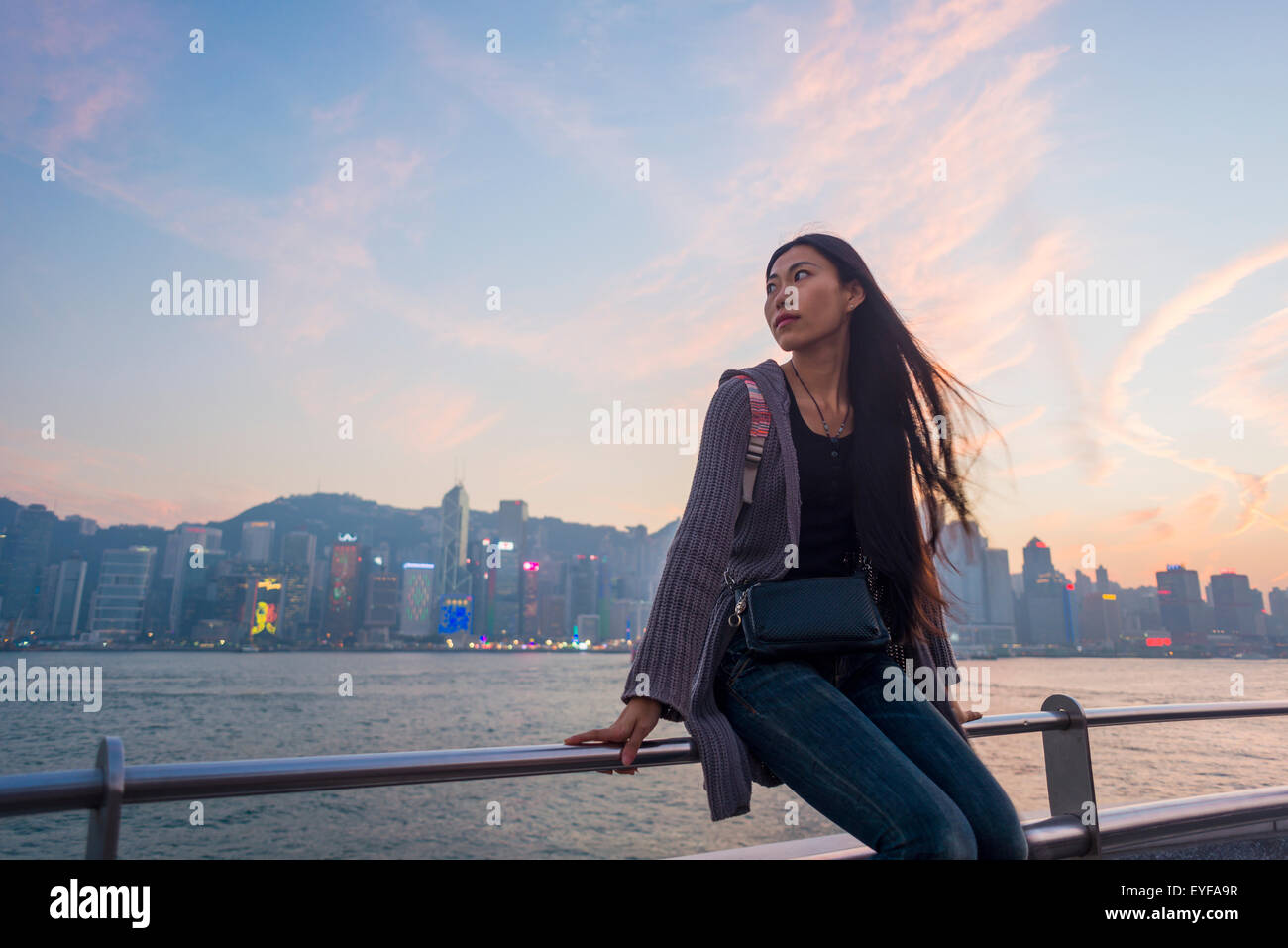 Une jeune femme au bord de l'eau assis sur la rambarde au coucher du soleil avec l'horizon à l'arrière-plan, Kowloon, Hong Kong, Chine Banque D'Images