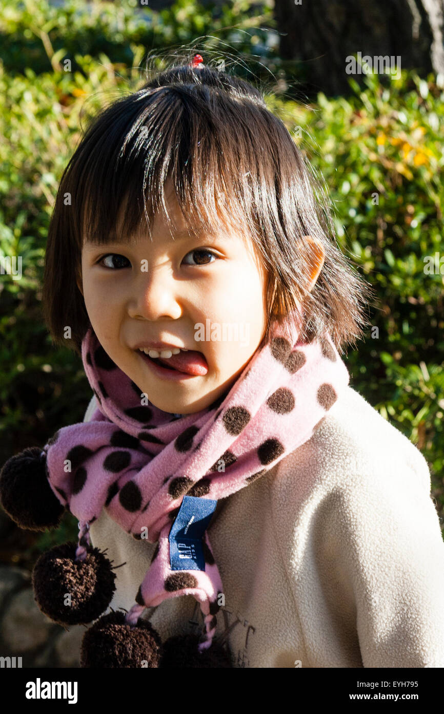 Japanese girl, tournant la tête pour regarder au spectateur, de la langue lolling out. Enfant, 4-5 ans. Porte manteau et écharpe. L'hiver. Close up, la tête et l'épaule. Banque D'Images