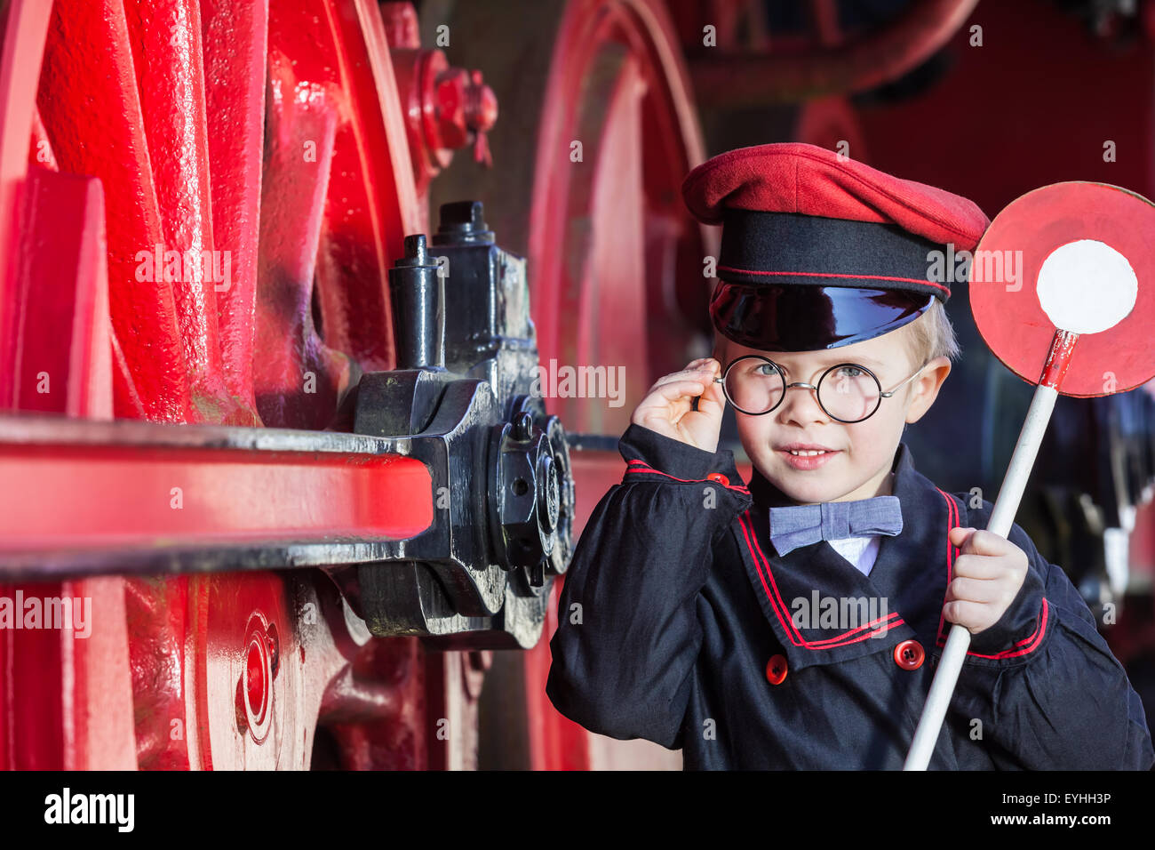 Petit enfant garçon comme chef de train nostalgique avec cap et disque de signalisation à côté de grandes roues d'une locomotive à vapeur Banque D'Images