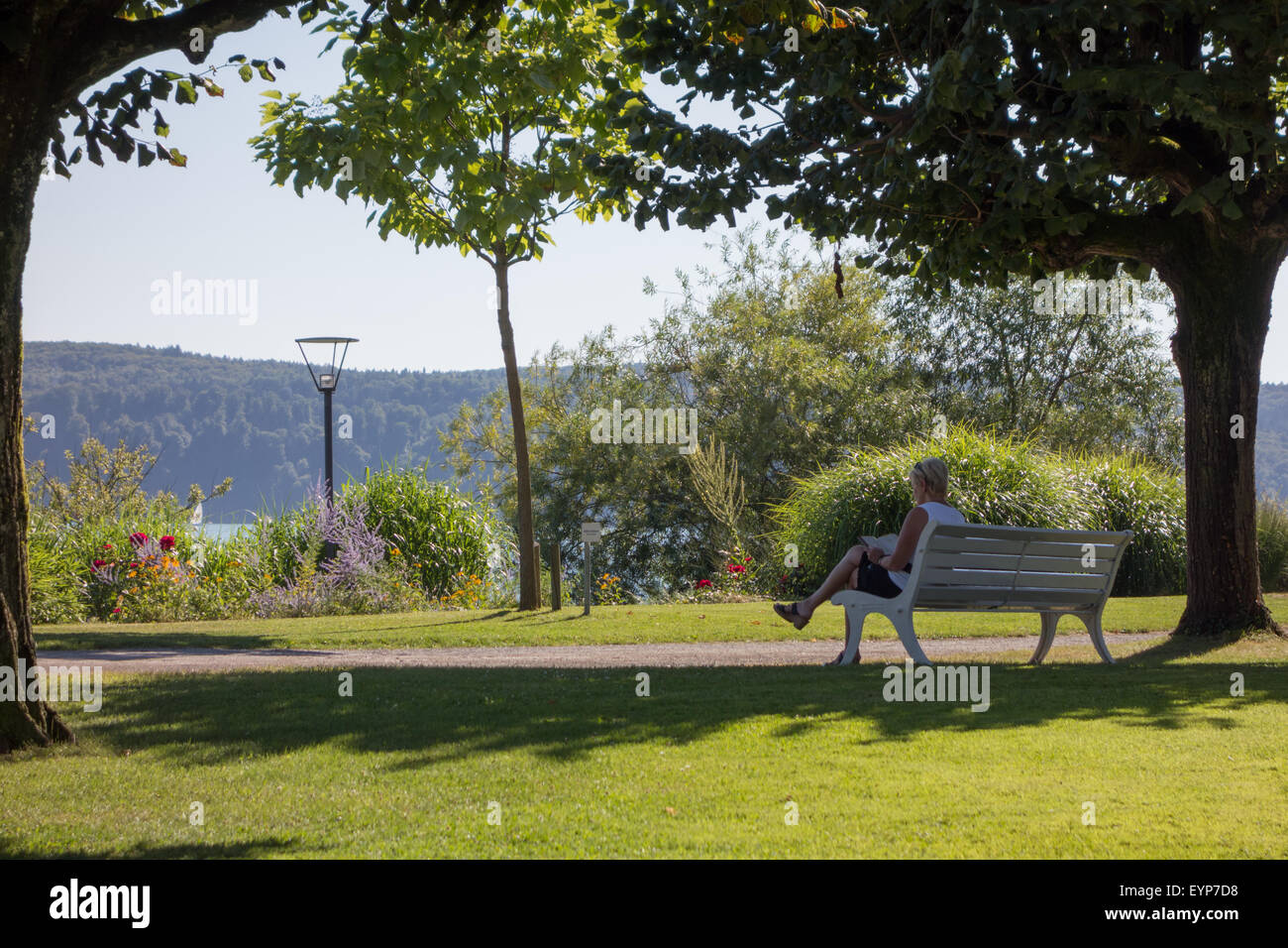 Une femme avec de courts cheveux blonds assise sur un banc à l'ombre d'un arbre pour lire un livre dans des jardins par le bord d'un lac Banque D'Images