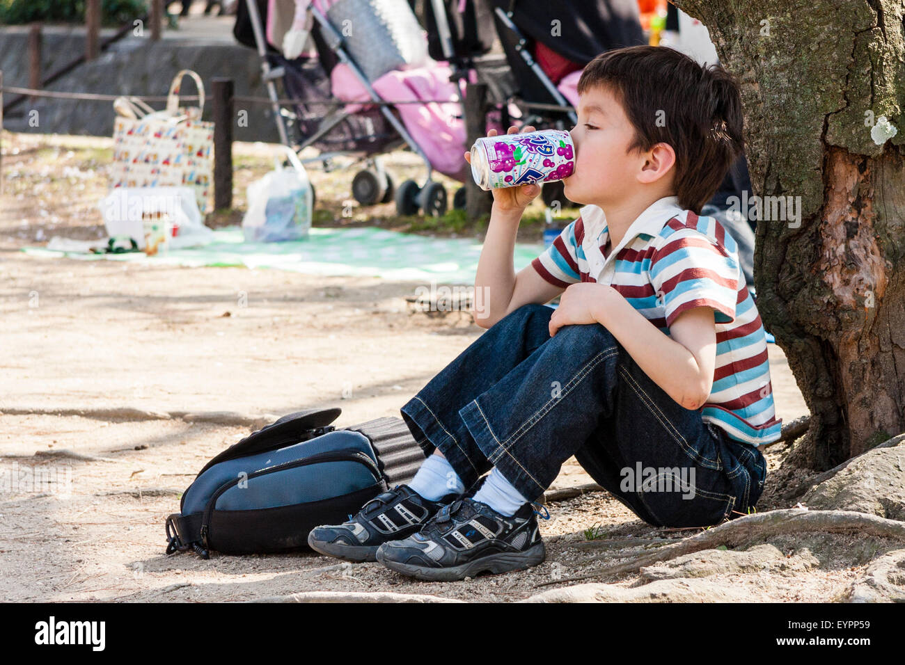 Caucasien enfant, garçon, 7-8 ans, assis à l'extérieur avec retour à l'arbre dans un parc public, de boire un verre de raisin japonais peuvent. Banque D'Images