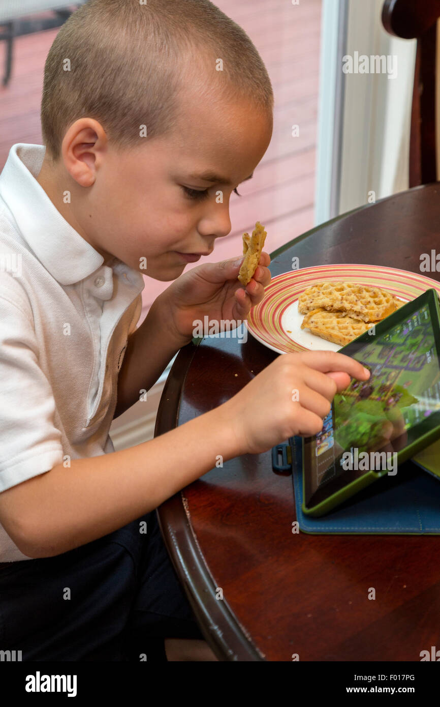 Jeune garçon (7 ans) Joue une partie sur son iPad en mangeant le petit déjeuner. M. Banque D'Images