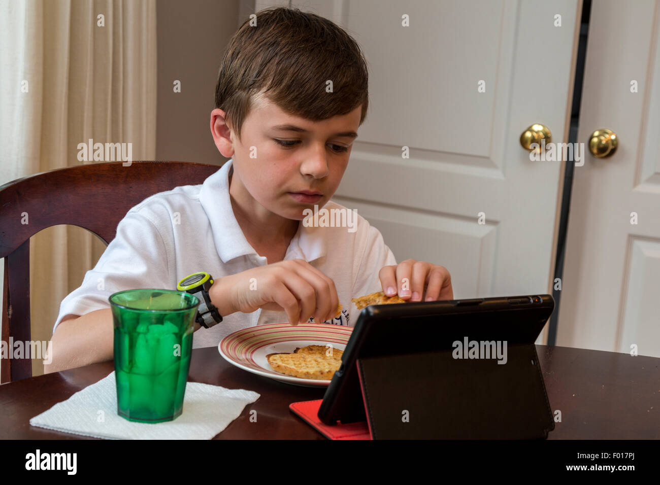 Jeune garçon (9 ans) regardant son iPad tout en prenant un petit déjeuner.M. Banque D'Images