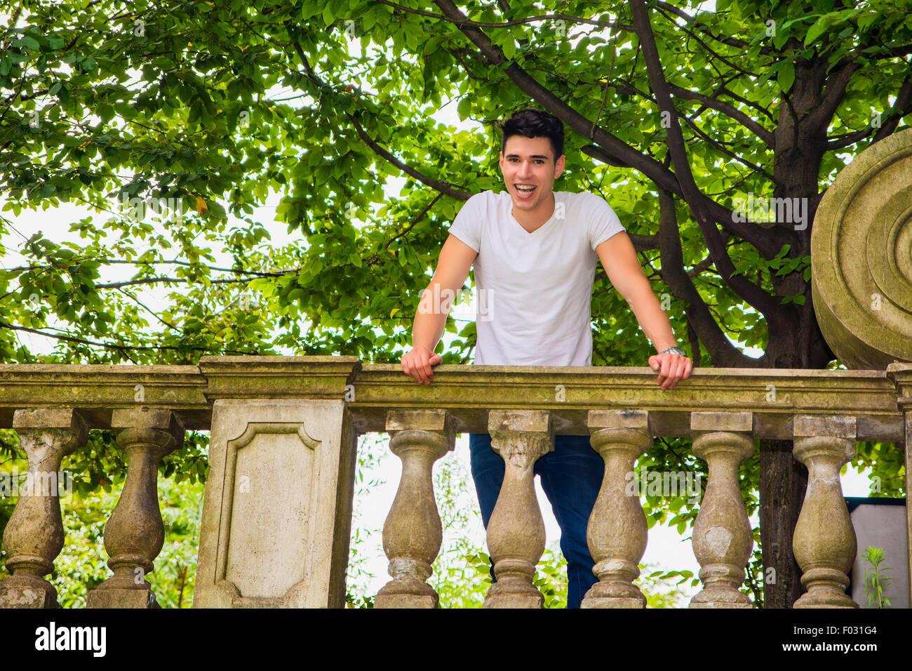 Beau jeune homme assis sur un balcon ou un pont s'appuyant sur la balustrade de pierre souriant à l'appareil photo Banque D'Images