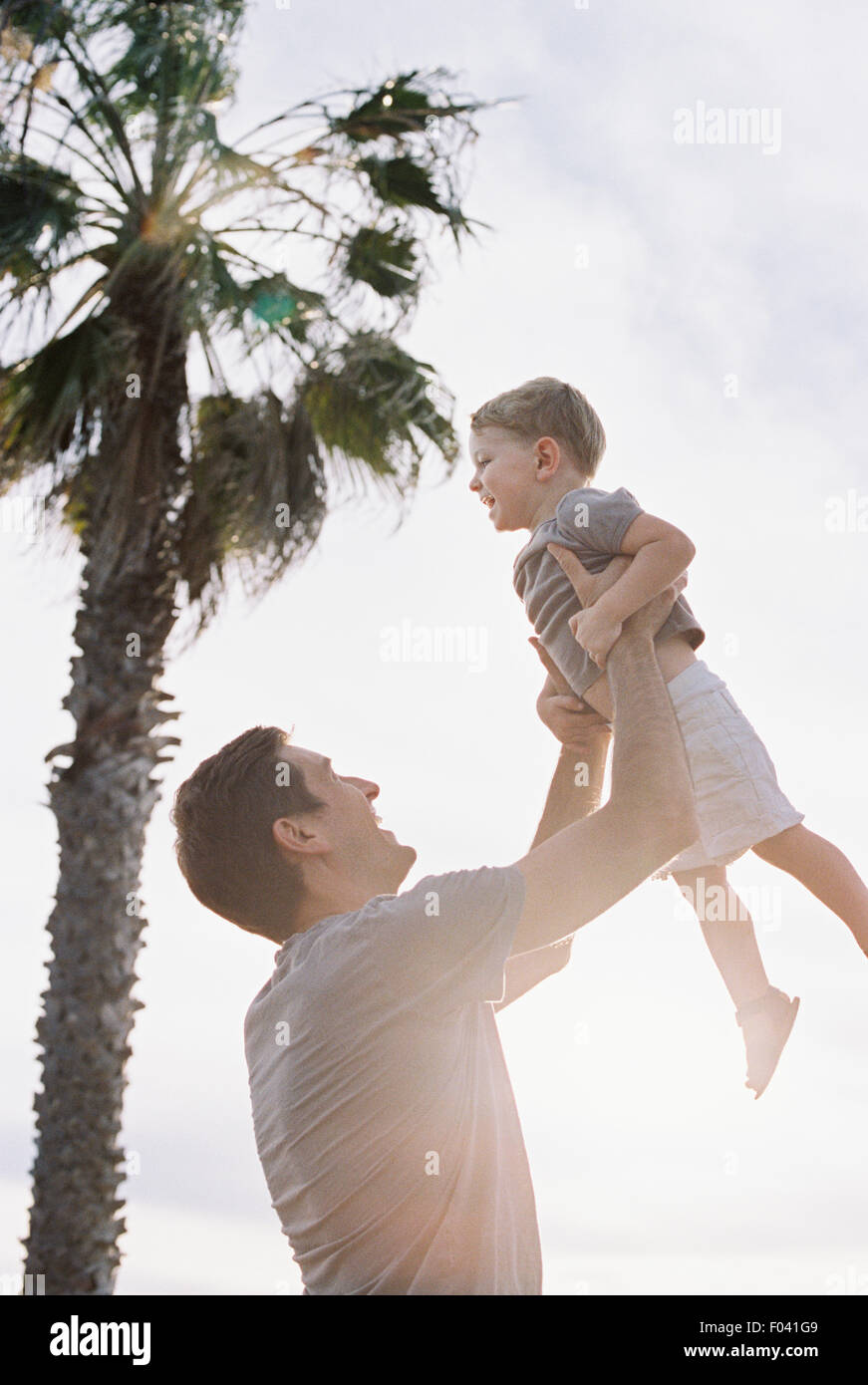 Homme debout par un palmier, jouant avec son jeune fils, le soulevant en l'air. Banque D'Images