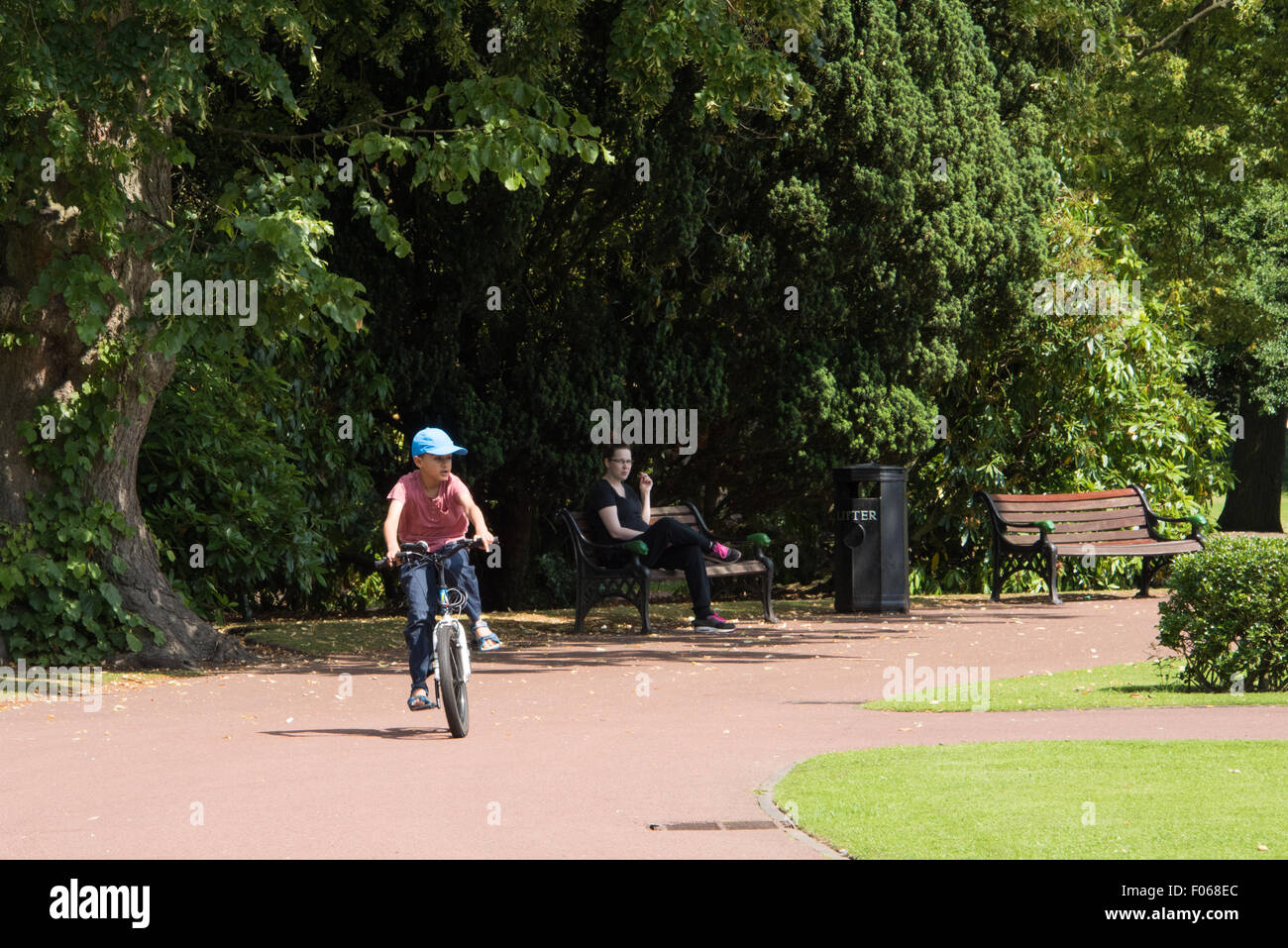 Un jeune garçon sur son vélo à travers le parc de l'Ouest avec une dame de le regarder à partir d'un banc à Wolverhampton West Midlands UK Banque D'Images