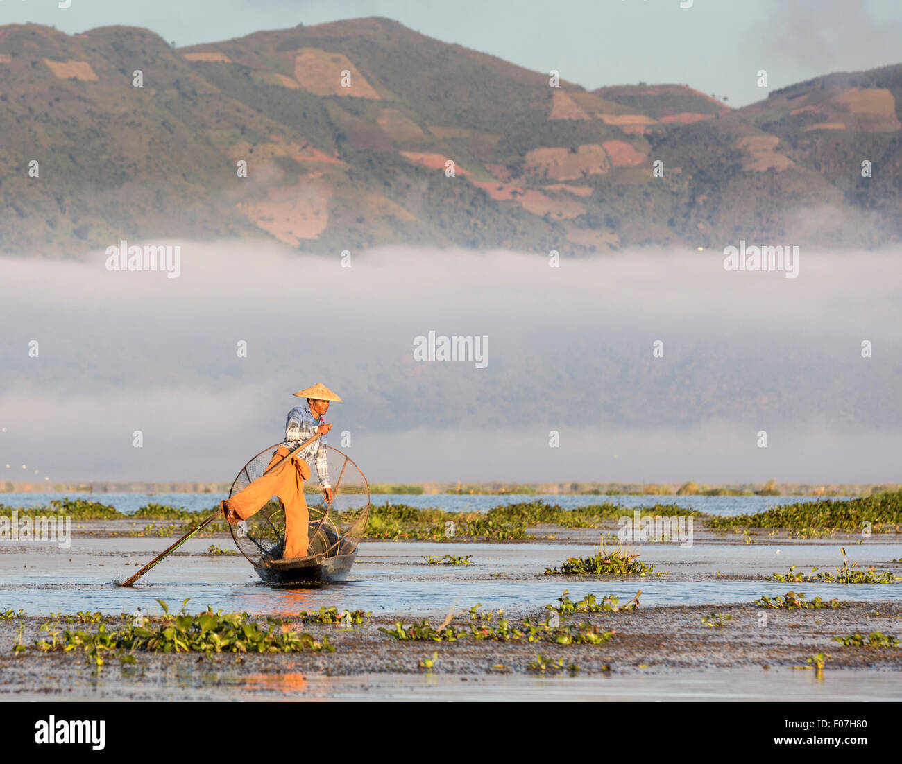 À l'aide d'un pêcheur du lac Inle à tenir la jambe dans la mode typique de oar. Morning Mist suaires montagnes derrière. Banque D'Images