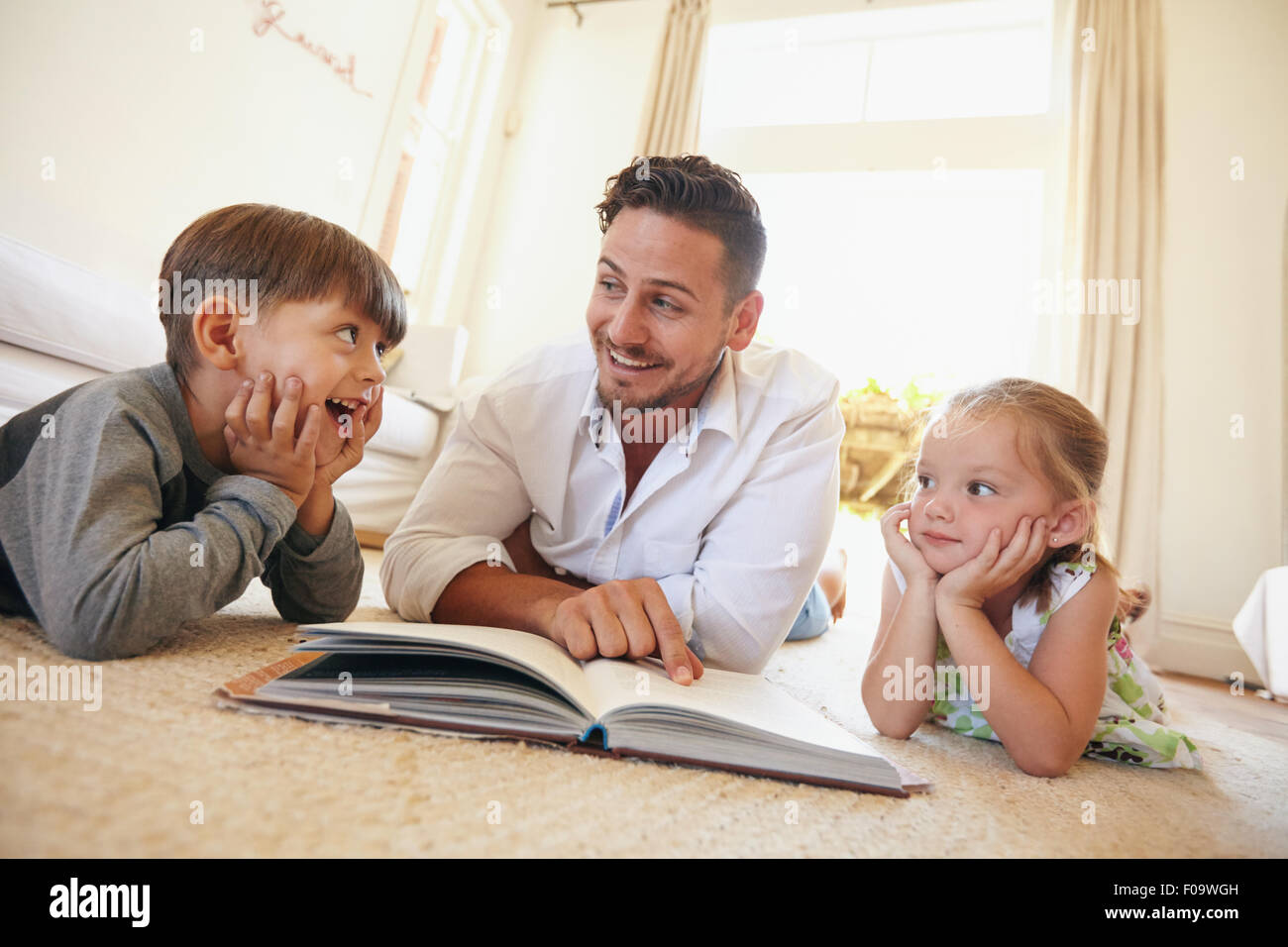 Photo de heureux jeune famille se trouvant sur le plancher de la lecture d'un livre. Jeune homme lisant des histoires à son petit-fils et sa fille à la maison. Banque D'Images