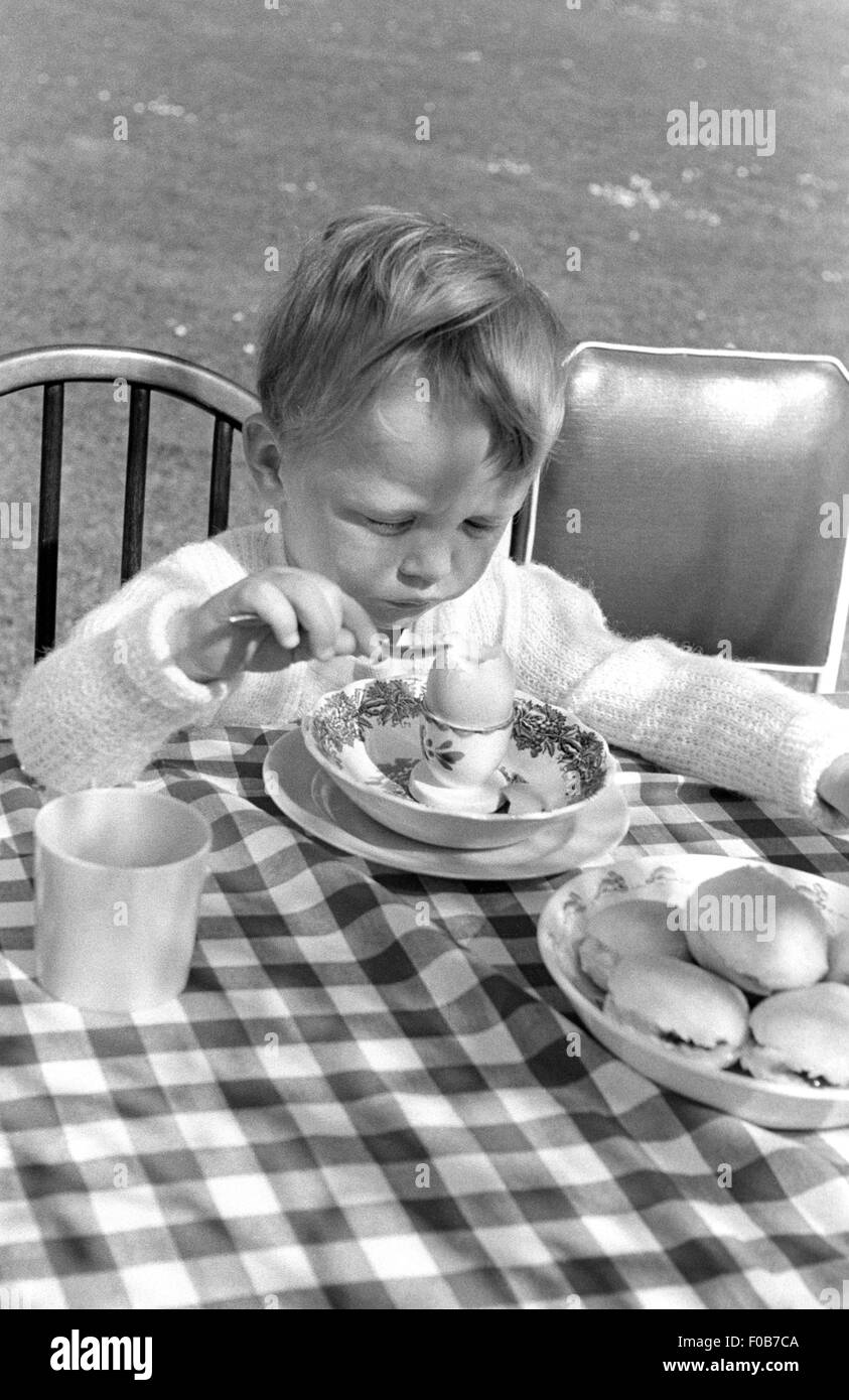 Un jeune garçon assis à la table en train de manger un œuf dur dans le jardin. Banque D'Images