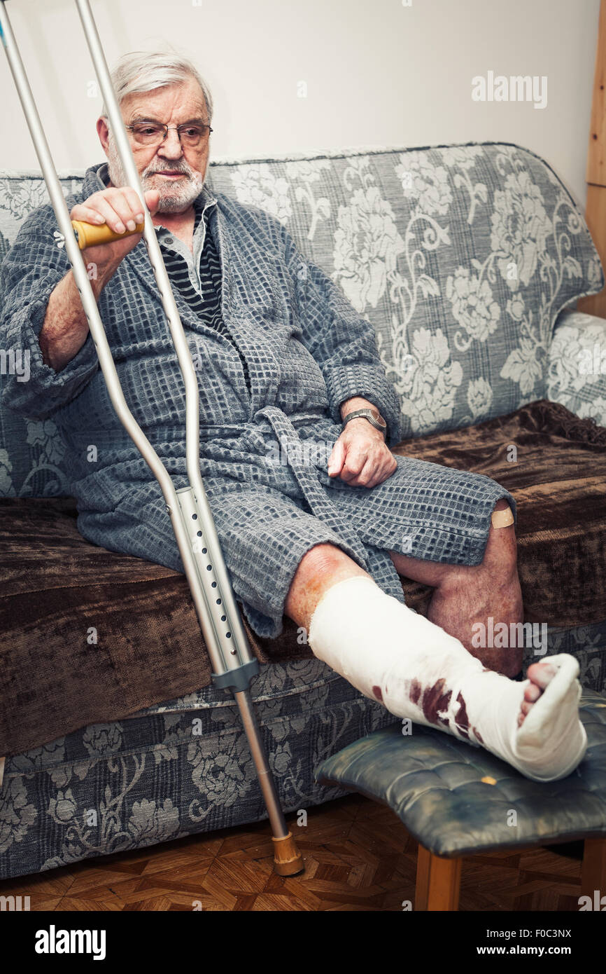 Senior man with broken leg sitting on sofa at home, tenant des béquilles Banque D'Images