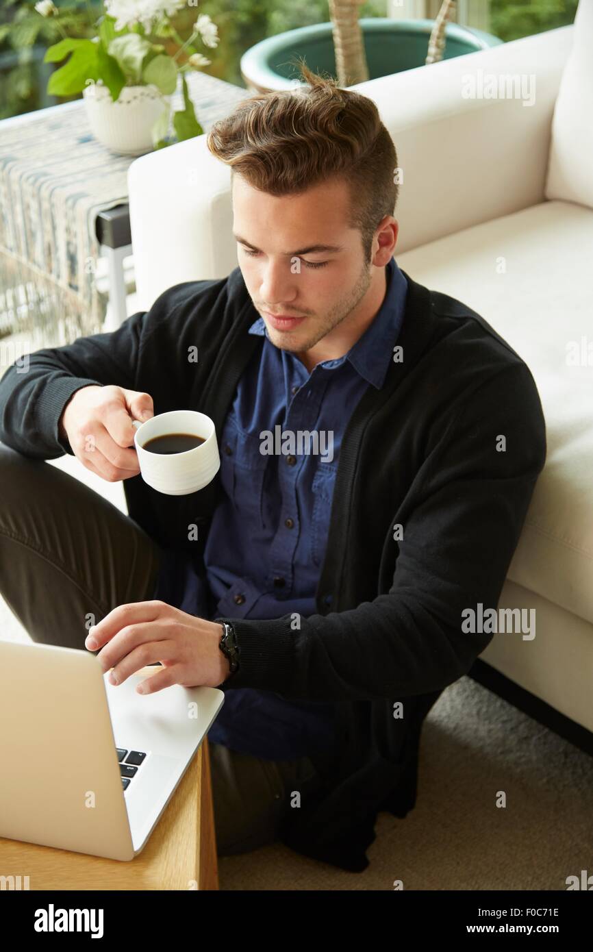 Portrait de l'homme assis sur le plancher à la maison looking at laptop et tasse de café potable Banque D'Images