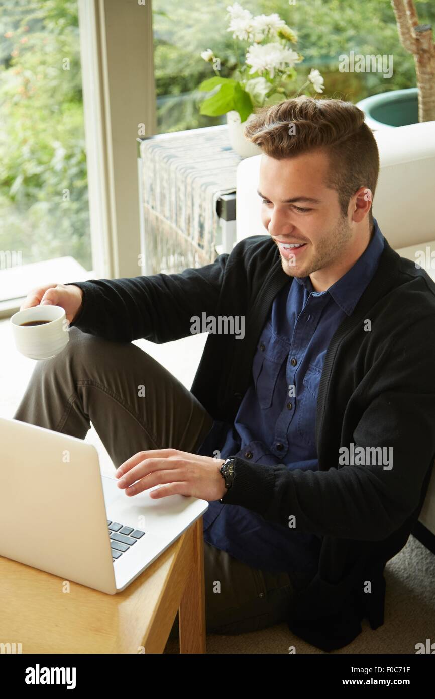 Portrait of smiling man sitting on floor at home looking at laptop et tasse de café potable Banque D'Images