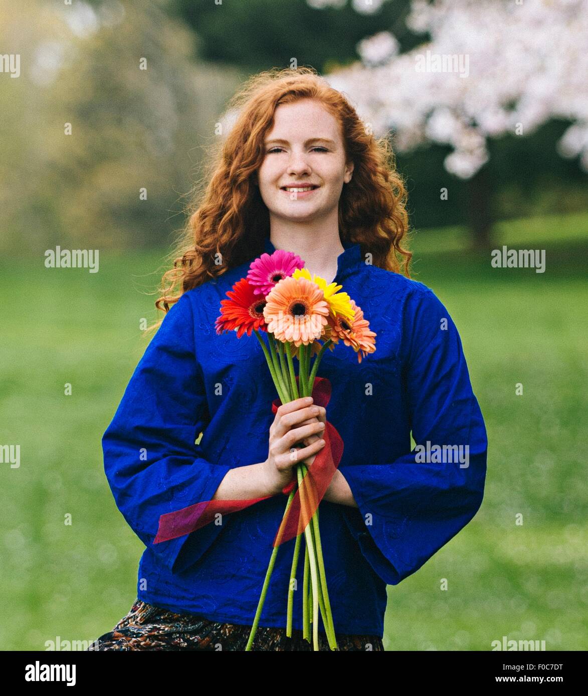 Portrait de jeune femme avec de longs cheveux roux ondulés holding bunch of flowers in park Banque D'Images
