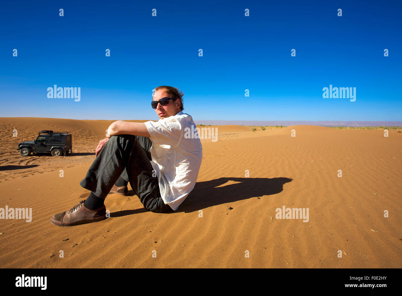Jeune homme assis sur une dune de sable à Merzouga et regardant la caméra, à l'arrière-plan, il y a un 4x4 véhicule stationné. Banque D'Images