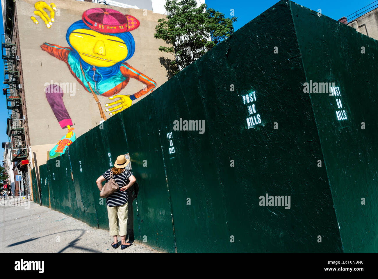 New York, NY - 14 août 2015 - Un passant à travers la clôture peeps pour regarder les artistes de rue lits jumeaux brésiliens Os Gemeos créer une murale de 6 étages, dans le quartier de l'East Village de Manhattan Banque D'Images