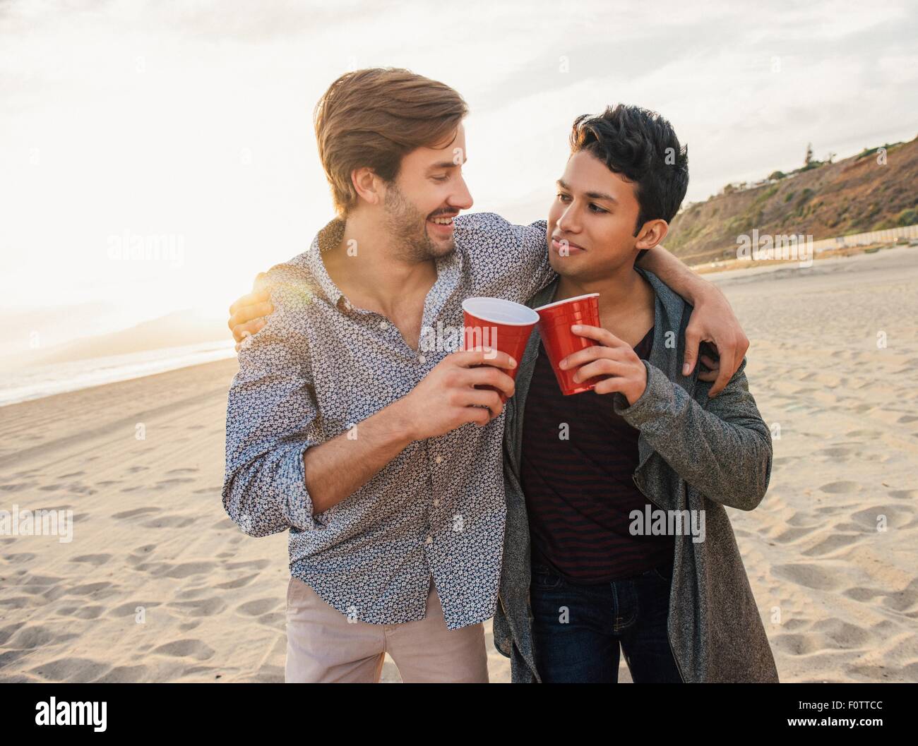 Deux amis de sexe masculin ayant un verre ensemble à la plage, les bras autour de l'autre Banque D'Images