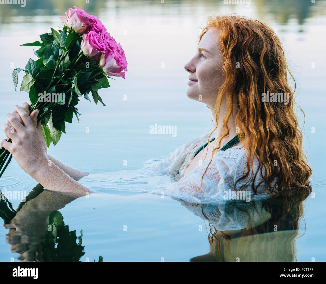 Tête et épaules de jeune femme aux longs cheveux rouges dans le lac en regardant bouquet de roses Banque D'Images