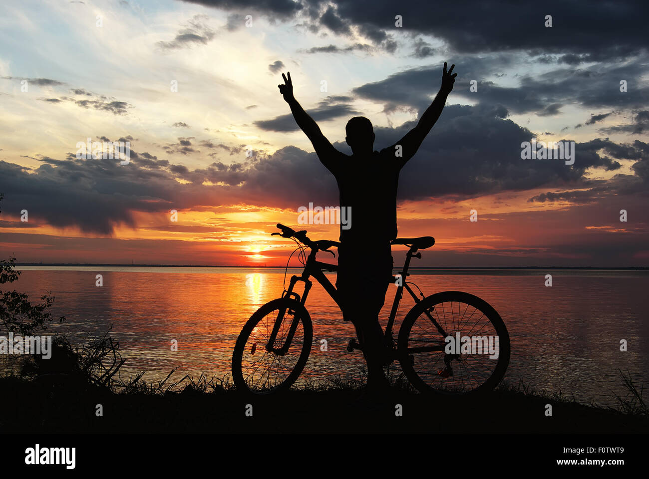 Silhouette d'un homme avec un vélo de montagne sur la rive du fleuve au coucher du soleil Banque D'Images