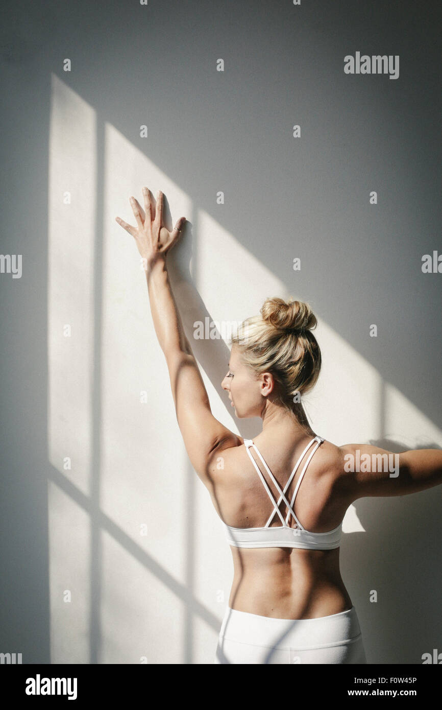 Une femme blonde, debout devant un mur blanc, faire du yoga, son bras levé, en touchant le mur. Banque D'Images