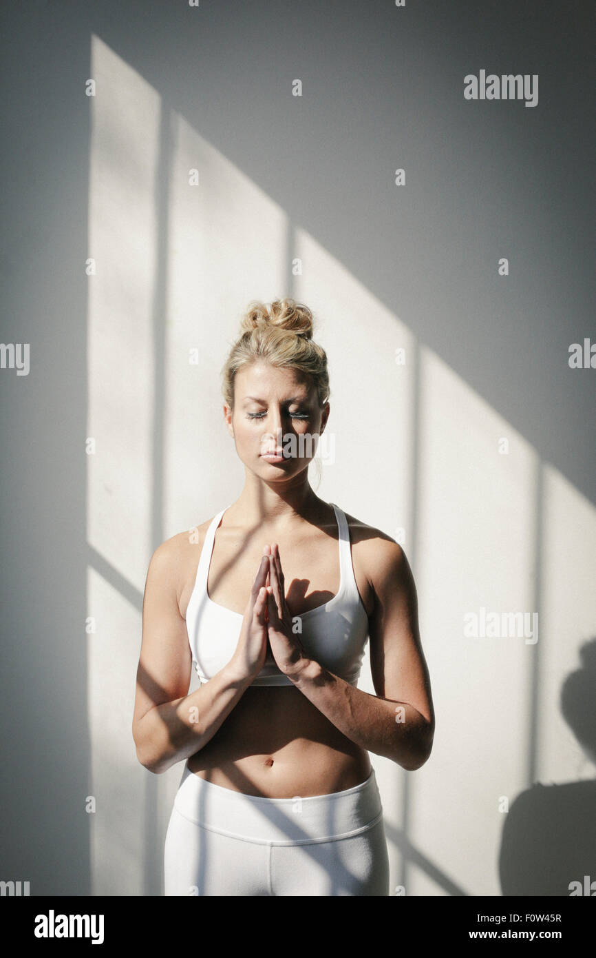 Une femme blonde, les yeux fermés, dans un blanc petit top et leggings, debout devant un mur blanc, faire du yoga. Banque D'Images