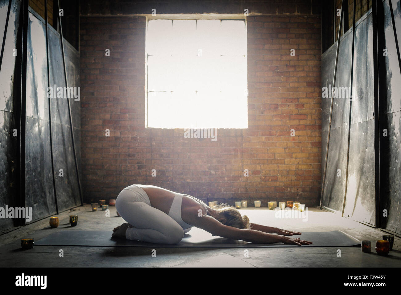 Une femme blonde, dans un blanc petit top et leggings,allongé sur un tapis de yoga sur le sol entourée de bougies, les bras tendus et s'accroupit. Banque D'Images