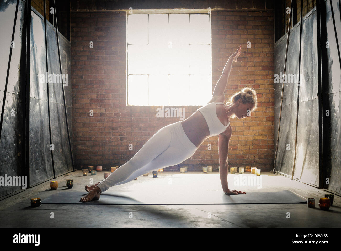 Une femme blonde, dans un blanc petit top et leggings, debout sur un tapis de yoga entouré par des bougies, faire du yoga, son bras levé. Banque D'Images