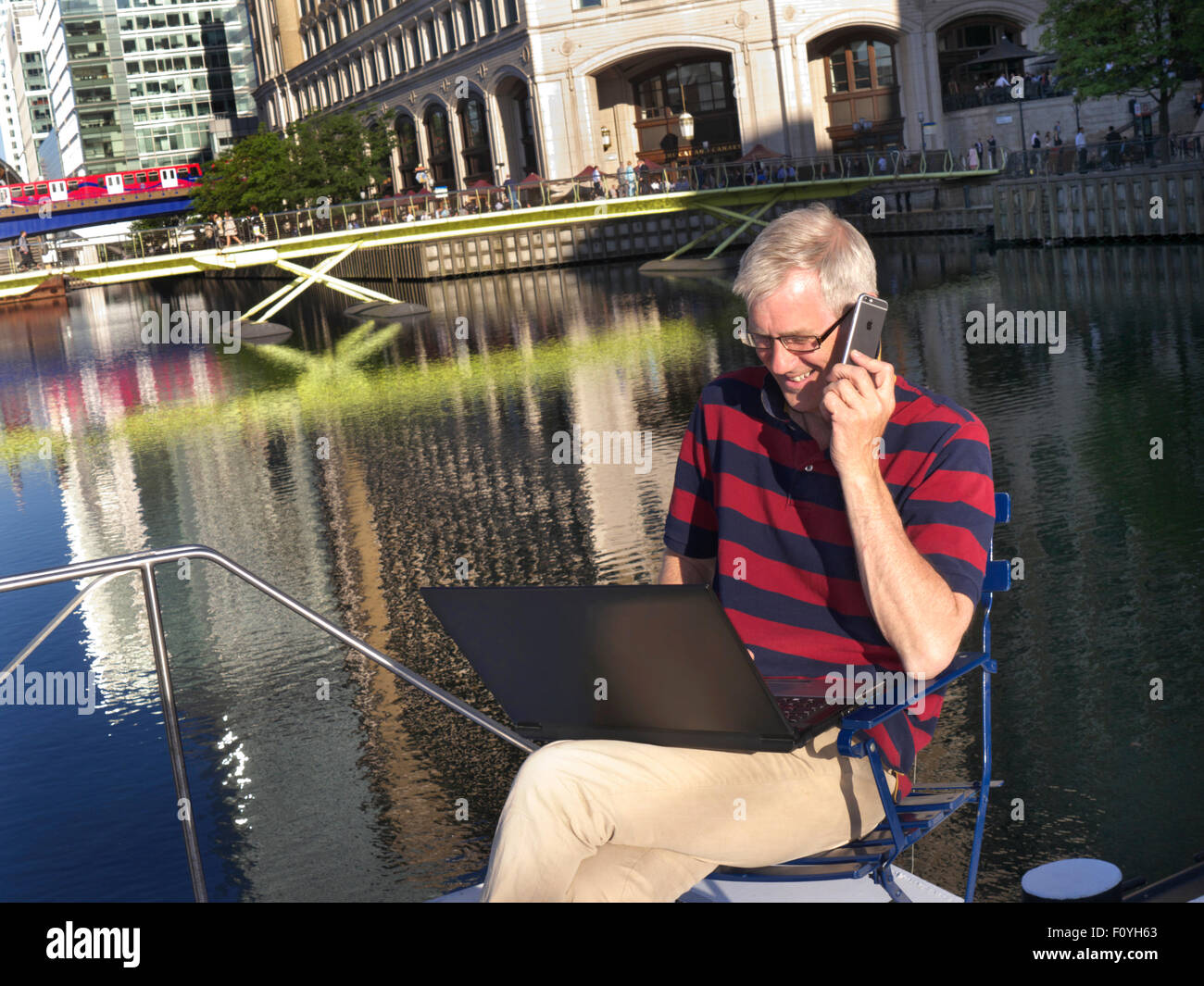 Homme mûr dehors sur son bateau en conversation sur iPhone 6 smartphone à son ordinateur portable derrière Canary Wharf Londres E14 Banque D'Images