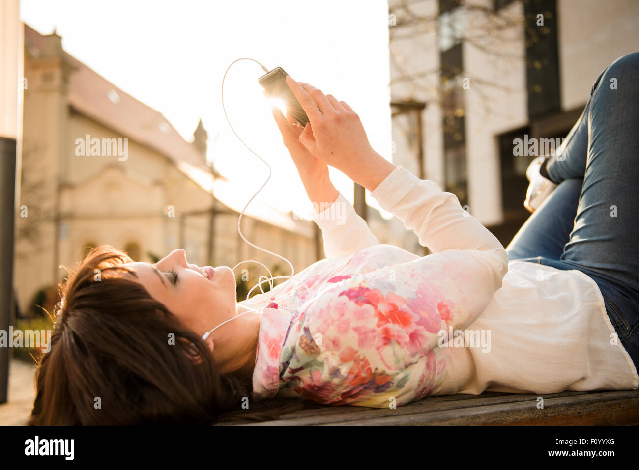 Young woman lying on écoute la musique de son banc téléphone dans street avec en arrière-plan Banque D'Images