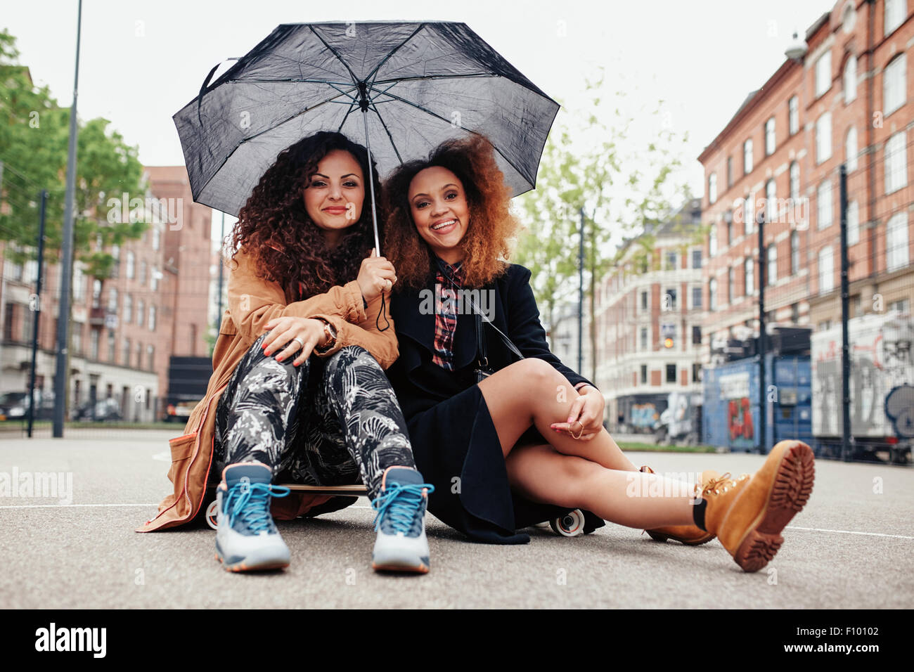 Deux jeunes femmes assises sur une planche à roulettes avec un parapluie. Femme amis assis ensemble sur rue avec skateboard à à c Banque D'Images