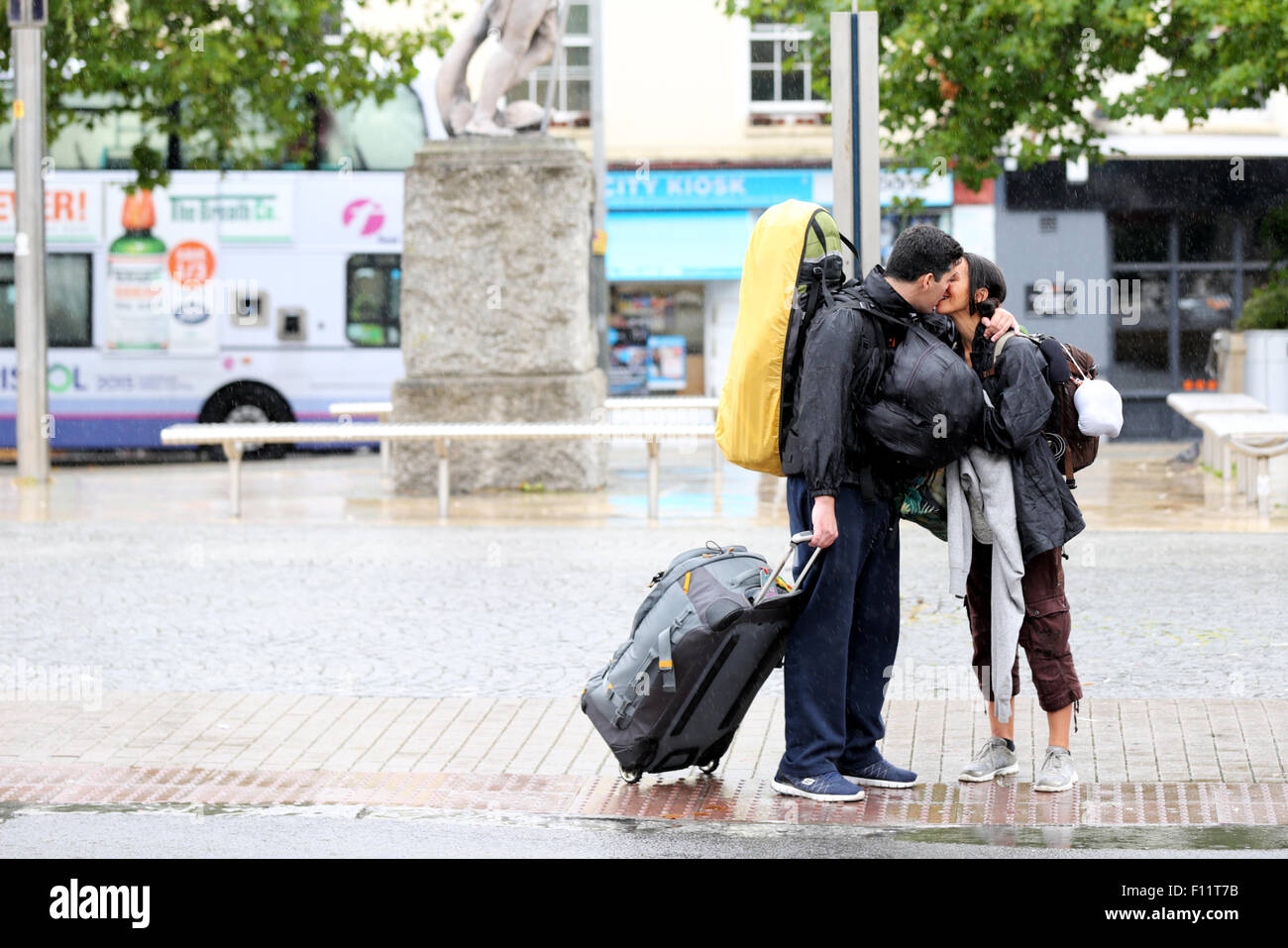 Un couple s'embrasser au revoir sur un après-midi pluvieux dans un centre-ville animé. Son versant avec la pluie mais le mâle a une valise et semble être la position off Banque D'Images