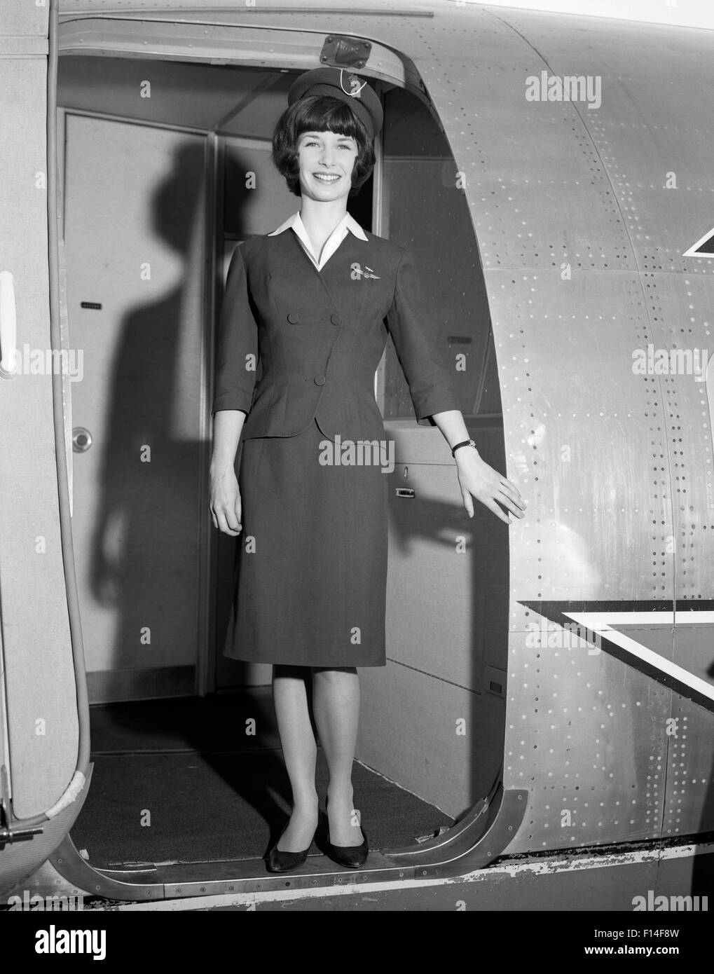 1960 SMILING STEWARDESS STANDING IN DOORWAY OF AIRPLANE LOOKING AT CAMERA Banque D'Images