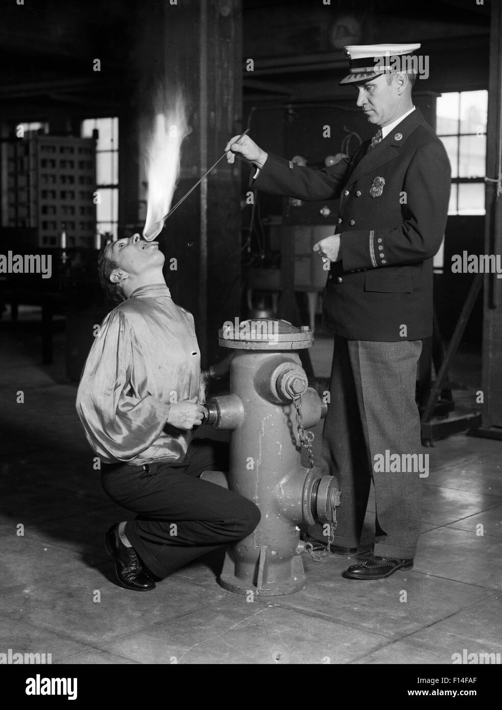 1930 Artiste de cirque homme mangeur de feu à genoux par poteau incendie avec l'homme ASSISTANT PORTANT BAGUETTE ENFLAMMÉE À BOUCHE Banque D'Images