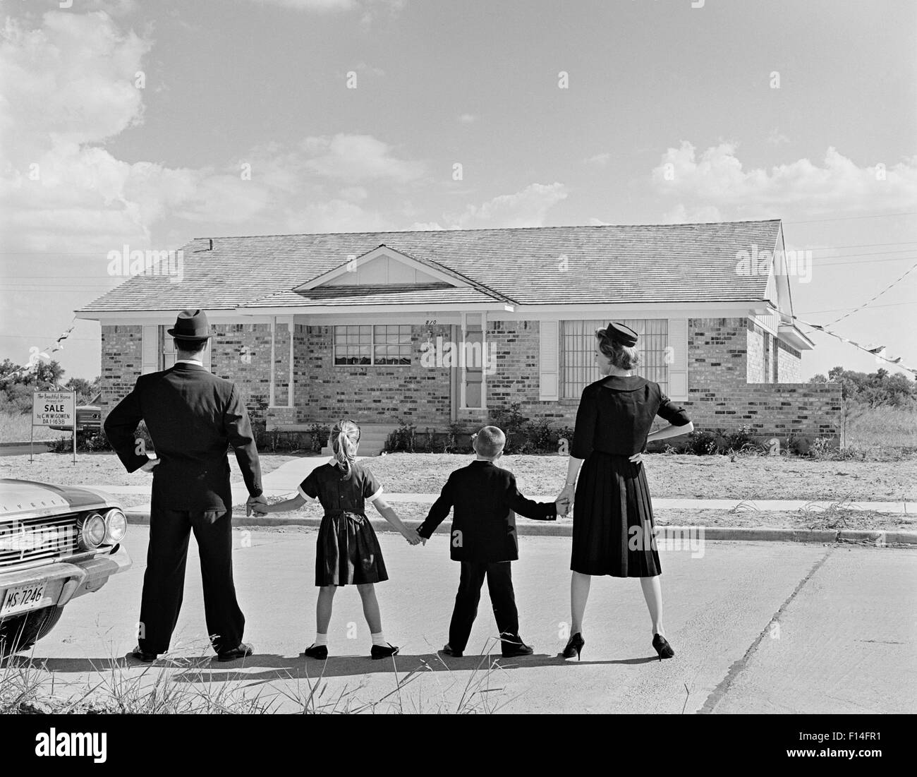 1960 FAMILLE DE QUATRE VUES DE DOS DEBOUT DEVANT DE NOUVEAUX SUBURBAN HOUSE HOLDING HANDS Banque D'Images