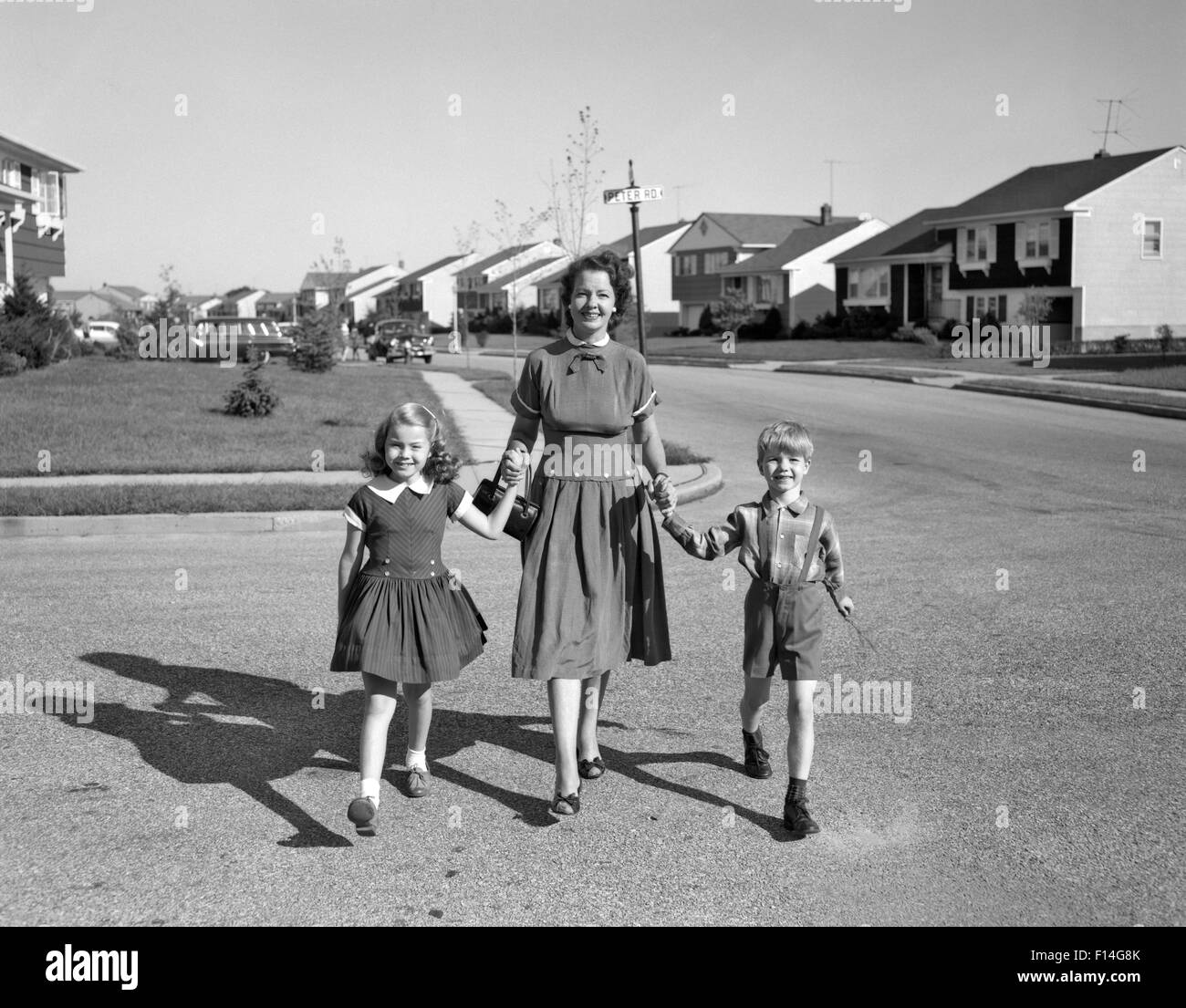 1950 SMILING HAPPY MOTHER HOLDING HANDS FILS ET FILLE DE MARCHER À TRAVERS LE DÉVELOPPEMENT DU LOGEMENT DE BANLIEUE STREET LOOKING AT CAMERA Banque D'Images