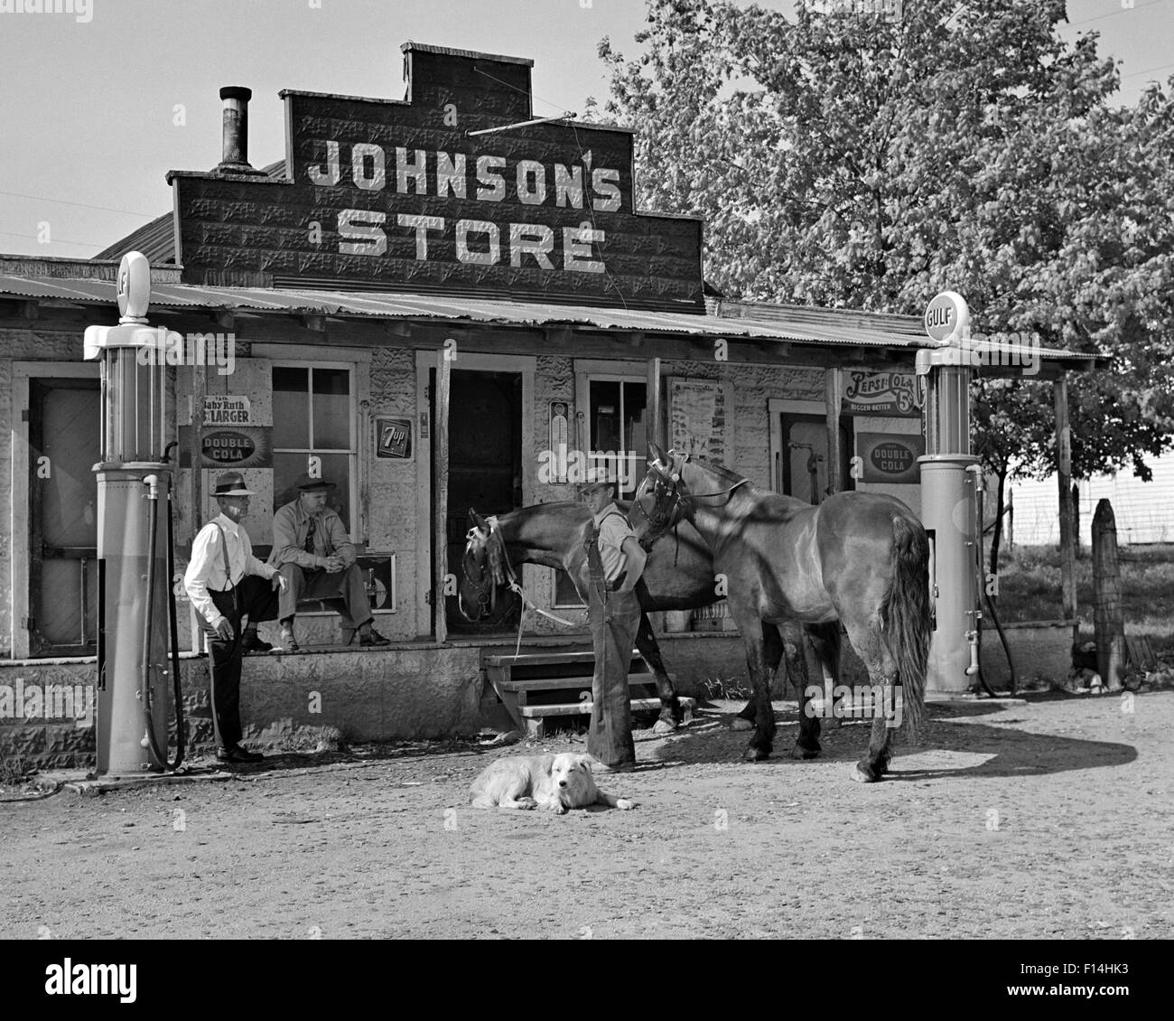 1930 GROUPE DE TROIS HOMMES DEUX CHEVAUX ET UN CHIEN À LA RECHERCHE SE SONT RÉUNIS DEVANT L'APPAREIL PHOTO AU PAYS DE L'ancien magasin CROCHETÉES PAR POMPES À ESSENCE Banque D'Images