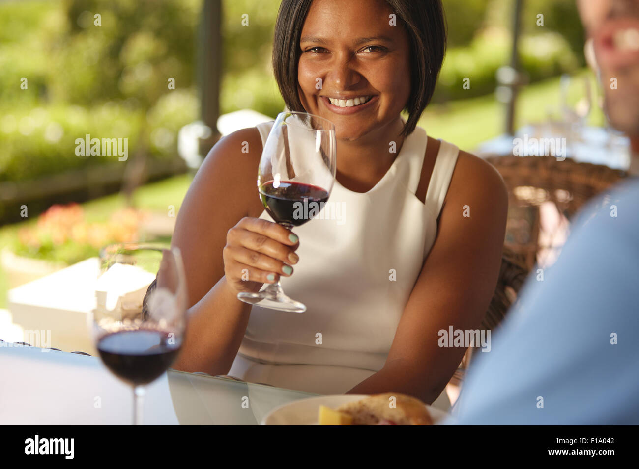 Portrait de belle jeune femme assise à une table en winery tenant un verre de vin rouge et souriant à la caméra. Banque D'Images