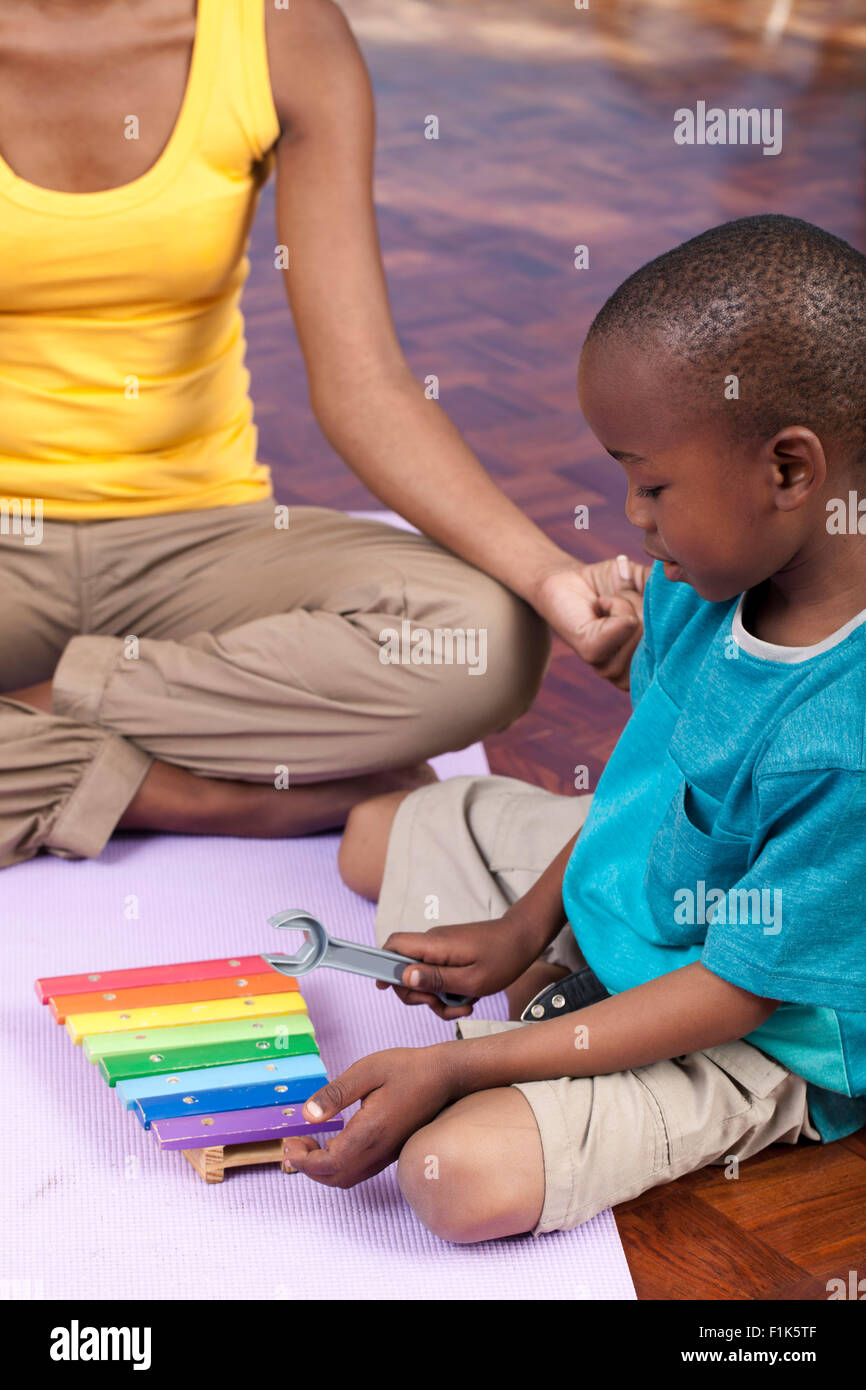 Jeune garçon africain joue avec un xylophone tandis que sa mère est assise en tailleur dans l'arrière-plan Banque D'Images